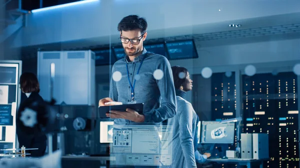 In Technology Research Facility: Chief Engineer Stands in the Middle of the Lab and Uses Tablet Computer (en inglés). Equipo de Ingenieros Industriales, Desarrolladores Trabajan en el Diseño del Motor Utilizan pizarra digital y — Foto de Stock