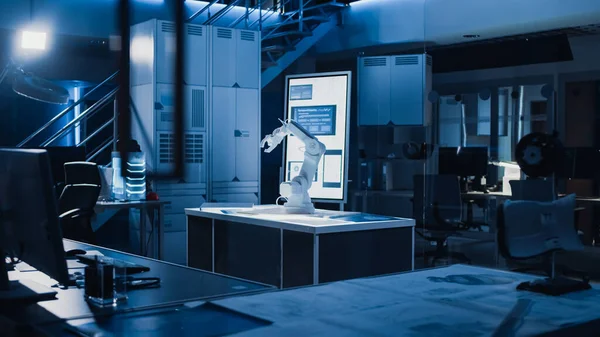 産業用ロボットデザイン研究室:机の上に立つロボットアームプロトタイプ。暗い背景の中で様々なハイテク機器、コンピュータ、デジタルホワイトボードとブループリント — ストック写真