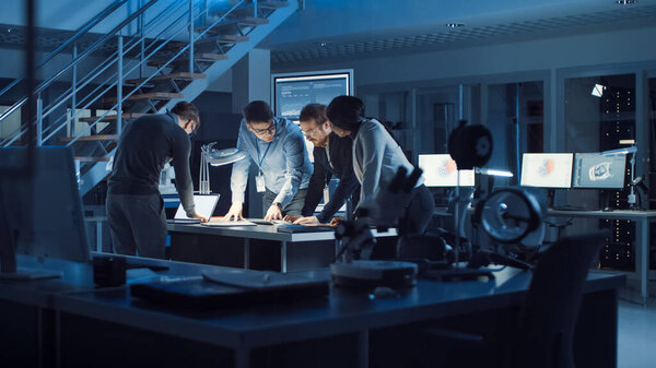 Разнообразная команда инженеров по разработке электроники, стоящая за рабочим столом, работающая с документами, решающая проблемы проекта поздно ночью. Специалисты, работающие над ультрасовременным промышленным дизайном.