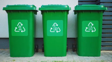 Yeşil renkli, plastik çöp kutuları, önünde farklı geri dönüşüm logoları olan, kahverengi ahşap duvara karşı sıra dizilmiş. Yiyecek, kağıt ve şişe için atık ayıklama kavramı. Ortam Çöp Kutusundan Kaydediliyor.