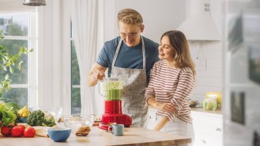 Gözlüklü yakışıklı genç adam önlük takıyor ve güzel kız mutfakta meyve püresi yapıyor. Mutlu çift sağlıklı organik içecek deniyor. Güneşli Bir Gün 'de Evde Erkek ve Kadın.