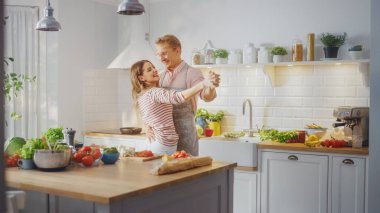 Pembe Gömlekli Yakışıklı Genç Adam ve Önlüklü Güzel Kız Mutfakta Yaratıcı Dans Ediyor. Sağlıklı Yeşil Sebzeli Sunny Modern Kitchen. Mutlu Çift Evde