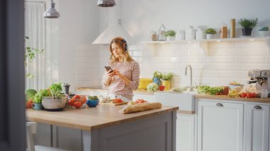 Modern Sunny Kitchen 'da Sağlıklı Organik Salata Yemekleri Hazırlayan Güzel Genç Kadın. Kulaklık takıyor ve bir cep telefonunda müzik kuruyor. Doğal Temiz Diyet ve Sağlıklı Yaşam Konsepti.