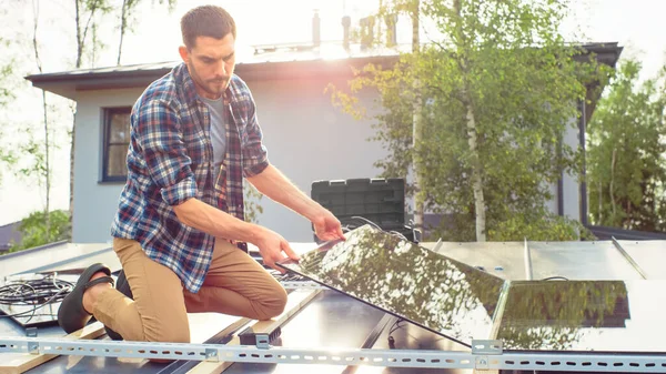 Biały mężczyzna w szachownicy instaluje czarne panele słoneczne na metalowej podstawie. Pracuje na dachu domu w słoneczny dzień. Koncepcja ekologicznej energii odnawialnej w domu. — Zdjęcie stockowe
