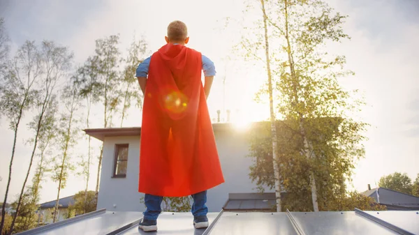 Pojken spelar rollen som superhjälte. Han står på taket till ett hus med händerna på midjan. Unge man bär en klar röd udde. Han tittar på solen.. — Stockfoto