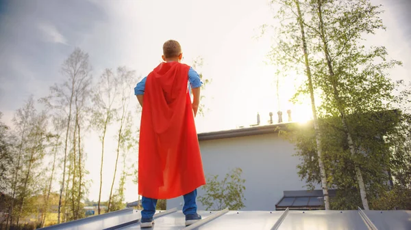 Pojken spelar rollen som superhjälte. Han står på taket till ett hus med händerna på midjan. Unge man bär en klar röd udde. Han tittar på solen.. — Stockfoto