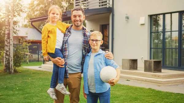 Portrait of a Happy Family of Three: Father, Daughter, Son. They Are Posing In Front of Camera on a Lawn Next to Their Country House and Smile. Dad is Holding the Girl in His Arms, Boy - Football. — Stock Photo, Image