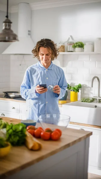 Счастливый молодой человек с длинным хаиром и синим пиджаком. Он прокручивает ленту новостей, социальные сети, чаты и сухой утренний кофе на кухне с здоровыми овощами. — стоковое фото