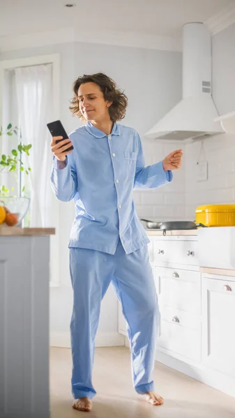 Happy Young Man with Long Hair Dancing at Home while Wearing Blue Pajamas. Poslouchá hudbu na mobilu. Energický muž pomocí chytrého telefonu v kuchyni se zdravou zeleninou. — Stock fotografie