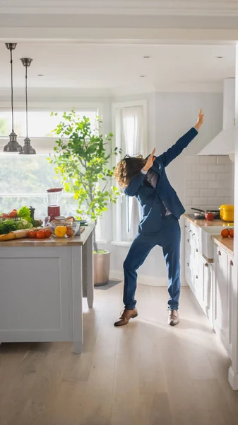 Ενεργητικός Αστείος Νεαρός με Μακριά Μαλλιά που ντύνεται στην Κουζίνα ενώ φοράει μπλε κοστούμι. Φωτεινό λευκό σύγχρονο χώρο κουζίνας με υγιή πράσινα λαχανικά σε ένα τραπέζι. Κάθετη λήψη ενός άνετου σπιτιού. — Φωτογραφία Αρχείου