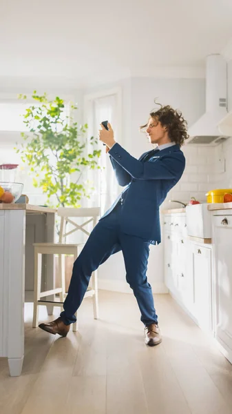 Счастливый молодой человек с длинными волосами танцует дома в синем деловом костюме. Он слушает музыку на мобильном телефоне. Энергичный человек с помощью смартфона на кухне со здоровыми овощами. — стоковое фото