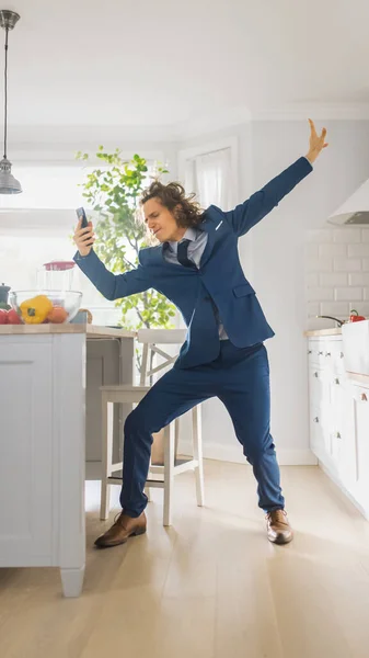Счастливый молодой человек с длинными волосами танцует дома в синем деловом костюме. Он слушает музыку на мобильном телефоне. Энергичный человек с помощью смартфона на кухне со здоровыми овощами. — стоковое фото
