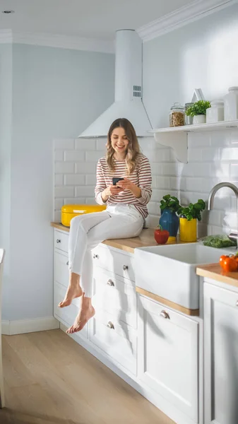 Красивая молодая женщина в полосатом джампере и белых штанах сидит на кухонной мебели и использует свой смартфон в современной солнечной кухне. Она счастлива и волнуется, просматривая Интернет. — стоковое фото