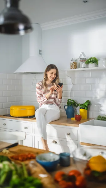 Krásná mladá žena v pruhované jumper a bílé kalhoty sedí na kuchyňském nábytku a pomocí svého chytrého telefonu moderní slunné kuchyně. Je šťastná a vzrušená při prohlížení internetu. — Stock fotografie