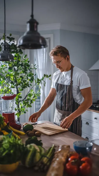 Jóképű férfi fehér ingben és kötényben egészséges bio salátát készít egy modern Sunny Kitchen-ben. Hipster Man szemüvegben főz. Természetes tiszta étrend és egészséges életmód koncepció. — Stock Fotó