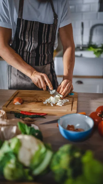 Schöner Mann schneidet mit einem scharfen Küchenmesser eine Frühlingszwiebel. Der Mann in weißem Hemd und Schürze bereitet in einer modernen, sonnigen Küche eine Bio-Salatmahlzeit zu. Natürliche saubere Ernährung und gesunder Lebensstil. — Stockfoto