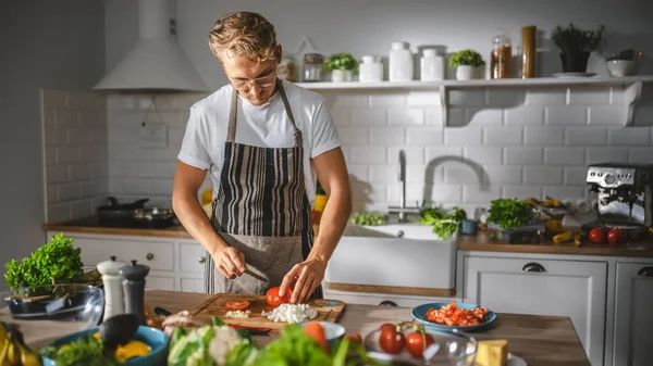 Le bel homme en chemise blanche et tablier prépare un repas de salade biologique sain dans une cuisine moderne ensoleillée. Hipster Man in Glasses Cooking. Concept d'une alimentation propre naturelle et d'un mode de vie sain. — Photo