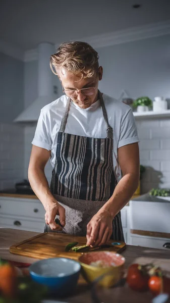Homem bonito em camisa branca e avental está fazendo uma refeição saudável salada orgânica em uma cozinha moderna ensolarada. Hipster Man in Glasses Cooking. Dieta Limpa Natural e Conceito de Modo de Vida Saudável. — Fotografia de Stock