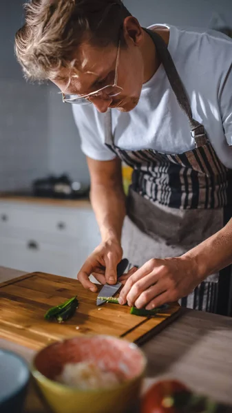 Bello uomo in camicia bianca e grembiule sta facendo un pasto sano insalata biologica in una cucina moderna soleggiata. Uomo hipster in cucina bicchieri. Dieta pulita naturale e concetto di stile di vita sano. — Foto Stock
