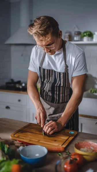 Schöner Mann in weißem Hemd und Schürze bereitet in einer modernen, sonnigen Küche eine gesunde Bio-Salatmahlzeit zu. Hipster Man in Brille beim Kochen. Natürliche saubere Ernährung und gesunder Lebensstil. — Stockfoto