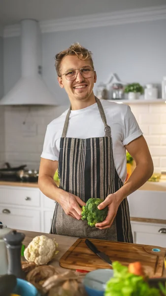 白いシャツとエプロンのハンサムな男は、現代のサニーキッチンで健康的な有機サラダ食事を作っています。メガネの中のヒップスター男はカメラで微笑む。自然食と健康的なライフスタイルのコンセプト. — ストック写真