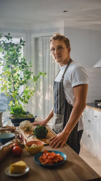 Schöner Mann in weißem Hemd und Schürze bereitet in einer modernen, sonnigen Küche eine gesunde Bio-Salatmahlzeit zu. Hipster-Mann mit Brille lächelt in die Kamera. Natürliche saubere Ernährung und gesunder Lebensstil. — Stockfoto