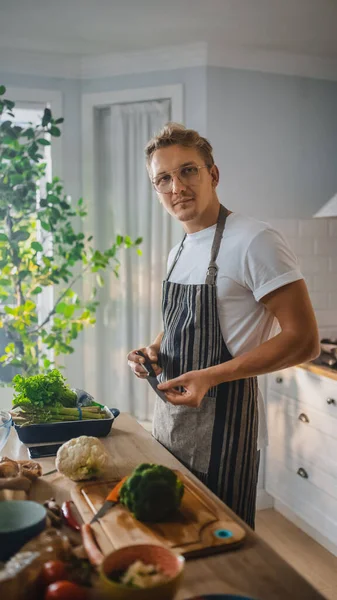 Schöner Mann in weißem Hemd und Schürze bereitet in einer modernen, sonnigen Küche eine gesunde Bio-Salatmahlzeit zu. Hipster-Mann mit Brille lächelt in die Kamera. Natürliche saubere Ernährung und gesunder Lebensstil. — Stockfoto