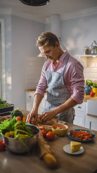 Schöner Mann in rosa Hemd und Schürze bereitet in einer modernen, sonnigen Küche eine gesunde Bio-Salatmahlzeit zu. Hipster-Mann mit Brille. Natürliche saubere Ernährung und gesunder Lebensstil. — Stockfoto