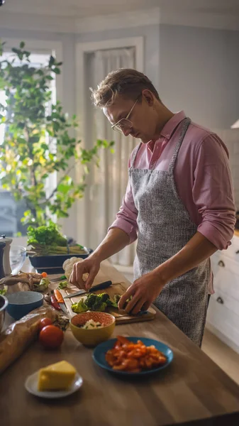 Schöner Mann in rosa Hemd und Schürze bereitet in einer modernen, sonnigen Küche eine gesunde Bio-Salatmahlzeit zu. Hipster-Mann mit Brille. Natürliche saubere Ernährung und gesunder Lebensstil. — Stockfoto