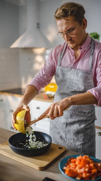 Handsome Man in Pink Shirt and Apron is Preparing a Healthy Vegetarian Meal on a Frying Pan. Healthy Organic Meal in a Modern Sunny Kitchen. Natural Clean Diet and Healthy Way of Life Concept.