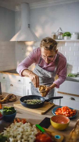 Bonito jovem preparando uma refeição vegetariana saudável em uma frigideira. Polvilhe alimentos verdes saudáveis com ervas e especiarias em uma cozinha moderna. Dieta Limpa Natural e Conceito de Modo de Vida Saudável. — Fotografia de Stock