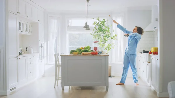 Nebun amuzant tânăr cu păr lung dansând în bucătărie în timp ce purta pijamale albastre. Bright White Modern Bucătărie zonă cu alimente verzi sănătoase pe o masă. Cozy Home . — Fotografie, imagine de stoc