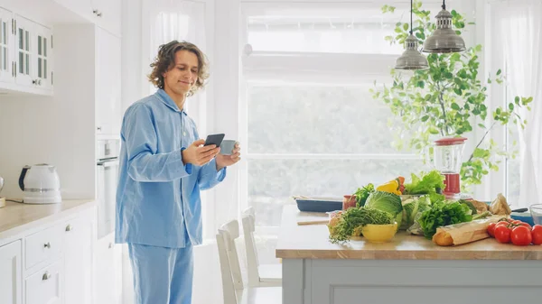 Счастливый молодой человек с длинным Хайром в китче и синей пижаме. Он является Scrolling Social Meadia и News Feed. Энергичный человек пьет кофе и здоровый завтрак. — стоковое фото