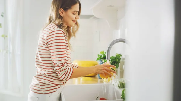 ストリップジャンパーの美しい若い女性は台所でトマトを洗っています。テーブルの上に健康的な緑の野菜と明るい白のモダンなキッチンエリア。Cozy Homeの幸せなカップル. — ストック写真