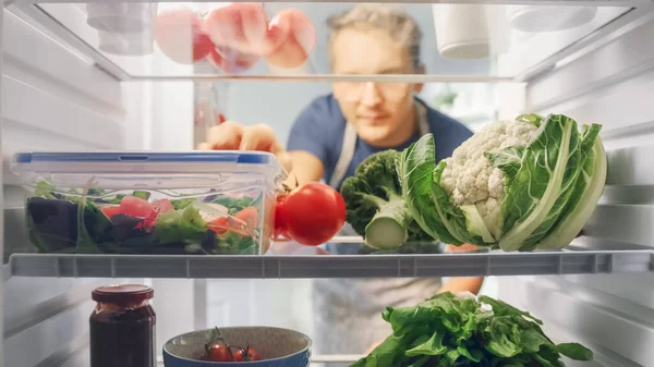 Guapo joven abre refrigerador lleno de alimentos orgánicos y agarra un montón de verduras para una ensalada. Concepto de dieta y estilo de vida saludable. POV Desde el interior del refrigerador. — Foto de Stock