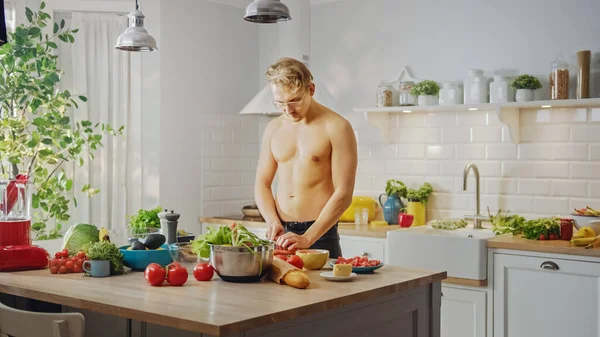 Bel homme sans chemise hacher une carotte avec un couteau de cuisine Sharp. Préparation d'un repas de salade bio sain dans une cuisine moderne. Mâle seins nus sur la diète. — Photo