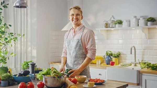 Schöner Mann in rosa Hemd und Schürze bereitet in einer modernen, sonnigen Küche eine gesunde Bio-Salatmahlzeit zu. Hipster-Mann mit Brille lächelt in die Kamera. Natürliche saubere Ernährung und gesunder Lebensstil. — Stockfoto