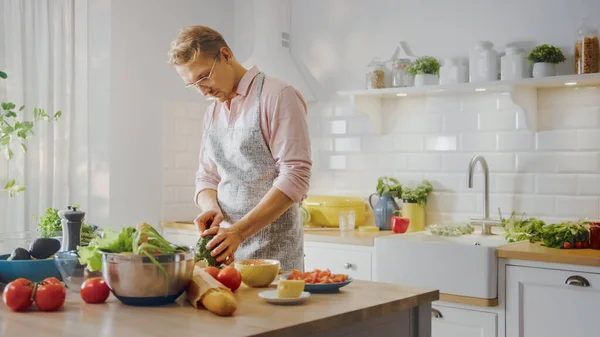 Hombre guapo con camisa rosa y delantal está haciendo una comida saludable ensalada orgánica en una cocina soleada moderna. Hombre Hipster con gafas. Concepto de dieta limpia natural y estilo de vida saludable. —  Fotos de Stock