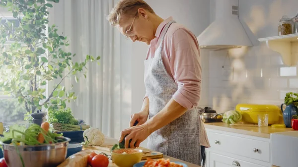 Hombre guapo con camisa rosa y delantal está haciendo una comida saludable ensalada orgánica en una cocina soleada moderna. Hombre Hipster con gafas. Concepto de dieta limpia natural y estilo de vida saludable. —  Fotos de Stock