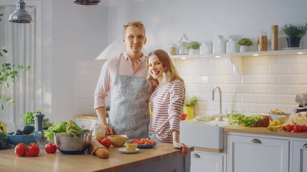 Красивый молодой человек в очках в фартуке и красивая девушка готовят салат на кухне. Счастливая пара обнимает друг друга. Естественный чистый рацион питания и здоровый образ жизни. — стоковое фото