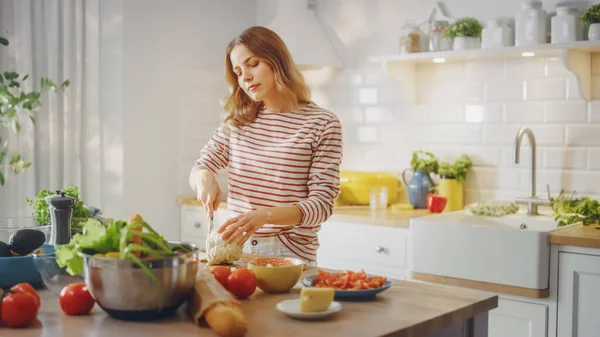 Молодая женщина в полосатом джампере делает здоровый органический салат на современной солнечной кухне. Настоящая женщина режет цветную капусту ножом. Естественный чистый рацион питания и здоровый образ жизни. — стоковое фото