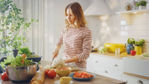 穿着条纹套头毛衣的年轻女性正在现代阳光厨房里做一个健康的有机沙拉。真正的女人是拿刀砍花的。自然清洁饮食与健康生活方式概念. — 图库照片