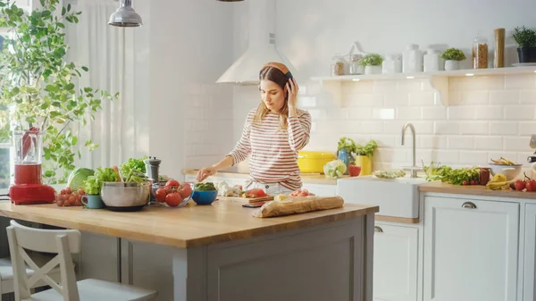 Красивая молодая женщина готовит здоровый органический салат на современной солнечной кухне. Она носит наушники и танцы под музыку с мобильного телефона. Естественный чистый рацион питания и здоровый образ жизни. — стоковое фото