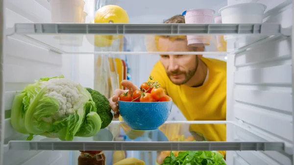 Réfrigérateur de cuisine intérieur : un bel homme prend des tomates cerises du réfrigérateur ouvert. Homme préparant un repas sain. Point de vue POV Tourné depuis un réfrigérateur rempli d'aliments sains — Photo
