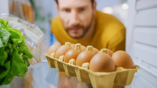 Schuss aus dem Inneren des Küchenkühlschranks: Schöner Mann öffnet Kühlschranktür, schaut hinein und denkt daran, Eier aus der Eierkiste zu holen. POV-Aufnahme aus dem Kühlschrank voller gesunder Lebensmittel — Stockfoto