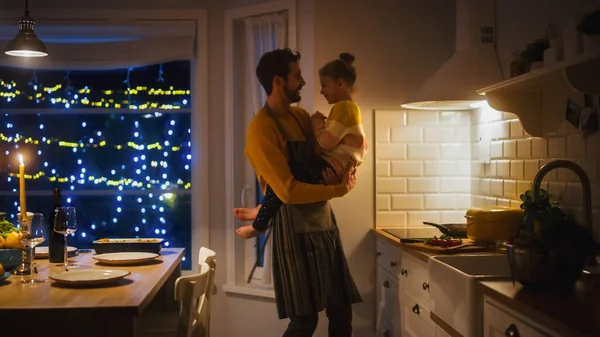 Isä ja tytär kokkaavat ja syövät yhdessä. Isä halaa söpöä pikkutyttöä. Juhlapöytä tyylikkäässä keittiön sisustuksessa lämpimällä valolla. — kuvapankkivalokuva