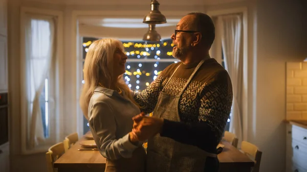 У старшей пары в любви есть романтический вечер, танцы на кухне, празднование годовщины. Счастливые пожилые муж и жена имеют прекрасный вечер с праздничным столом в уютной кухне. — стоковое фото