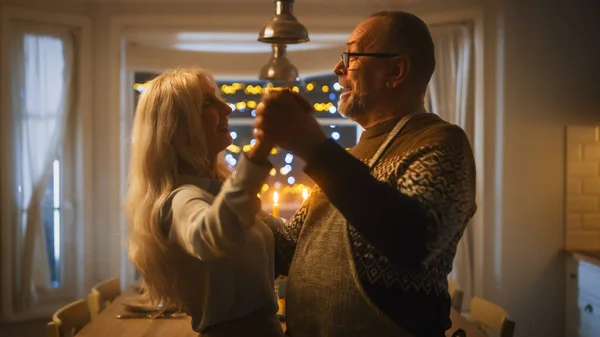 Pareja mayor enamorada tiene noche romántica, bailando en la cocina, celebrando el aniversario. Retrato de un feliz esposo y esposa ancianos tienen una velada encantadora con mesa festiva en la acogedora cocina — Foto de Stock