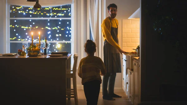 Gelukkige vader en dochter die samen koken en dineren. Vader bereidt eten, schattig klein meisje runt en knuffelt hem. Feestelijke tafel in stijlvol keukeninterieur met warm licht — Stockfoto