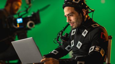 Handsome Smiling Actor Wearing Motion Capture Suit and Head Rig having Lunch Break, Sitting on Chair, Use Laptop. Studio High Budget Movie. On Film Studio Period Costume Drama Film Set clipart